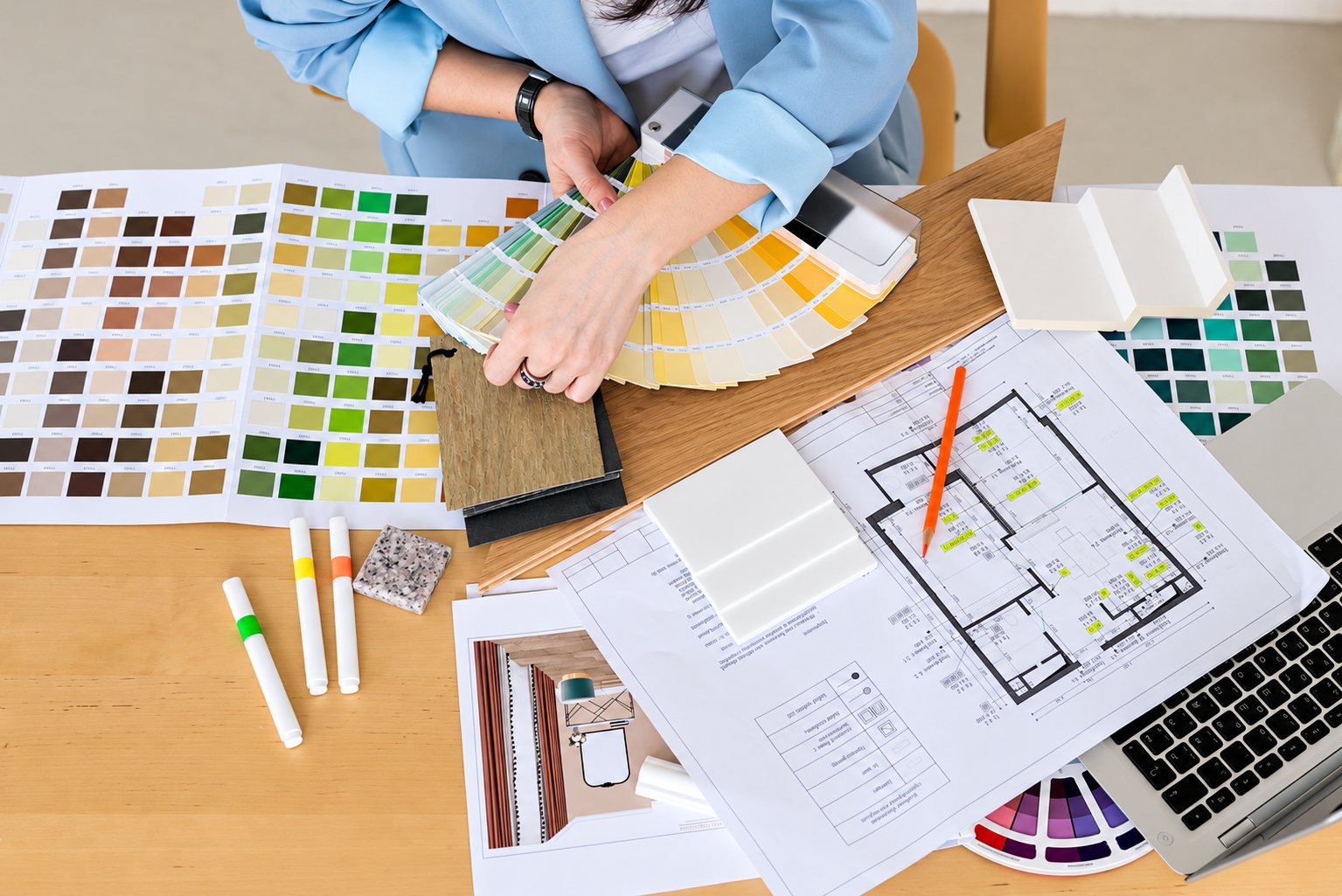 Vue du dessus d’une personne en chemise bleue qui tient un éventail de nuanciers de peinture, posé sur un bureau en bois avec plans, échantillons de matériaux, feutres et ordinateur portable.
