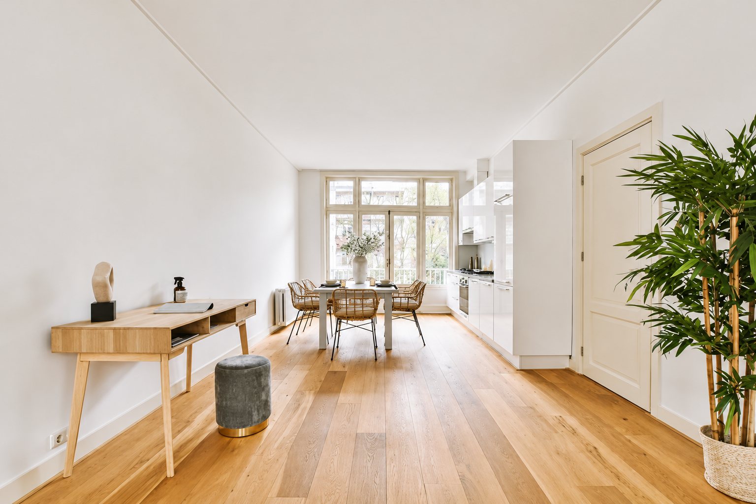 Pièce à vivre lumineuse au parquet clair, avec console en bois, pouf gris, table à manger près de la fenêtre et cuisine blanche.