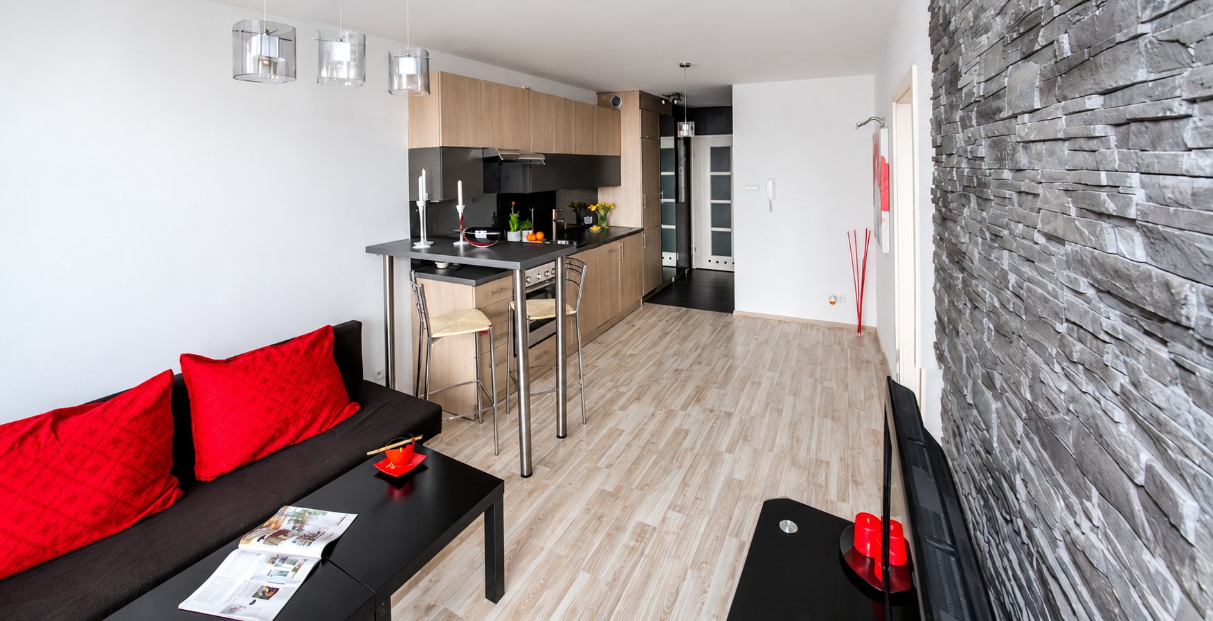Appartement lumineux avec canapé noir et coussins rouges, cuisine ouverte avec bar et mur de pierre grise.