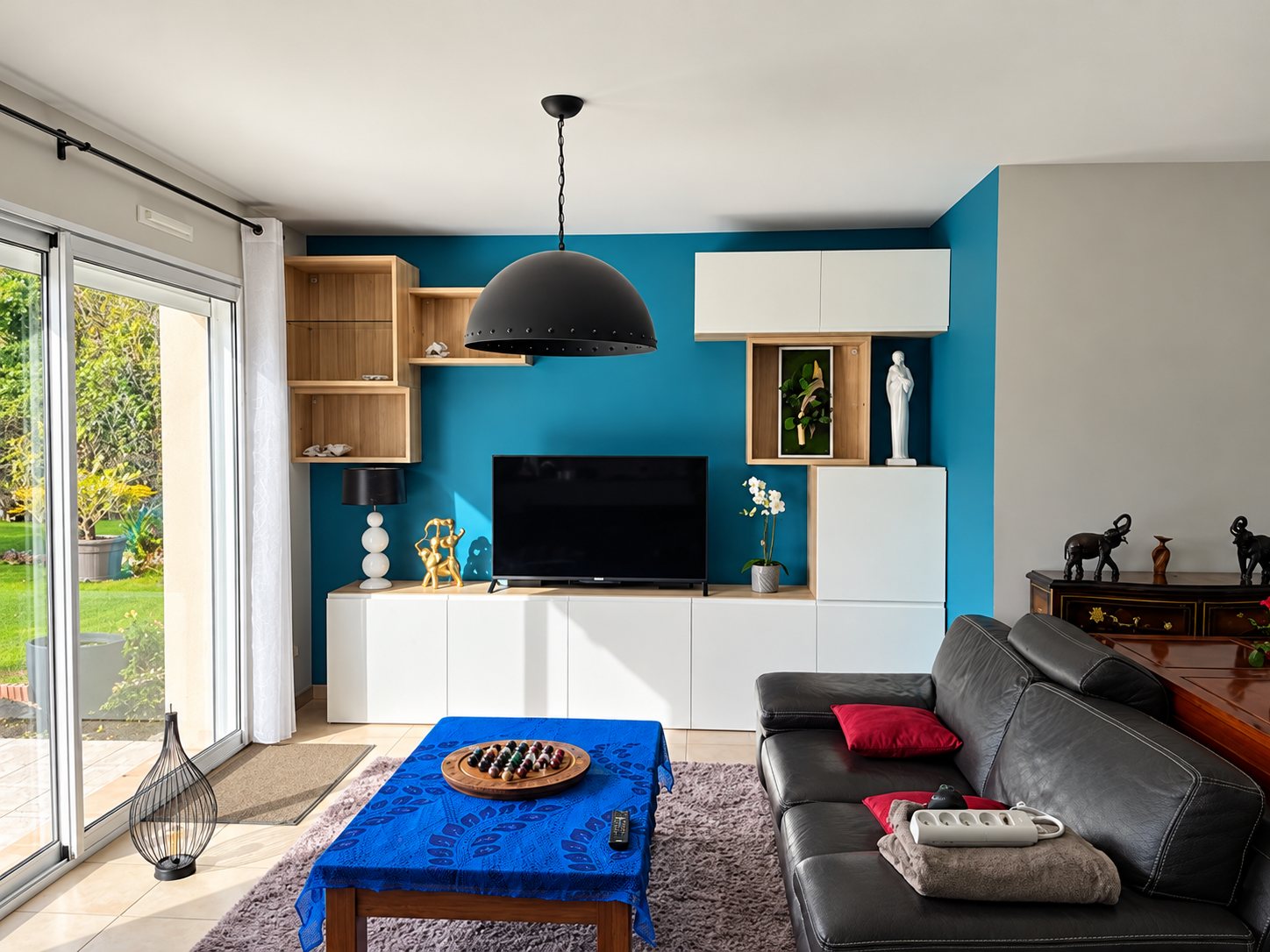 Un salon lumineux avec un mur bleu turquoise, un meuble bas blanc sous la télévision, une suspension noire, un canapé en cuir gris et une table basse recouverte d’un tissu bleu.