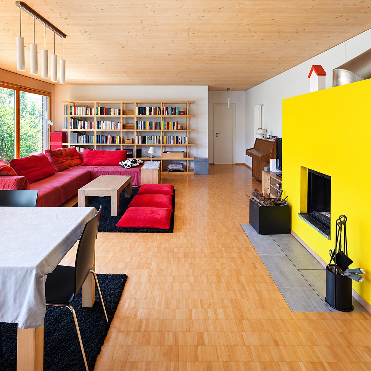Grand salon lumineux au plafond et parquet en bois clair, avec canapé rouge, bibliothèque, table à manger et cheminée encadrée d’un mur jaune vif.