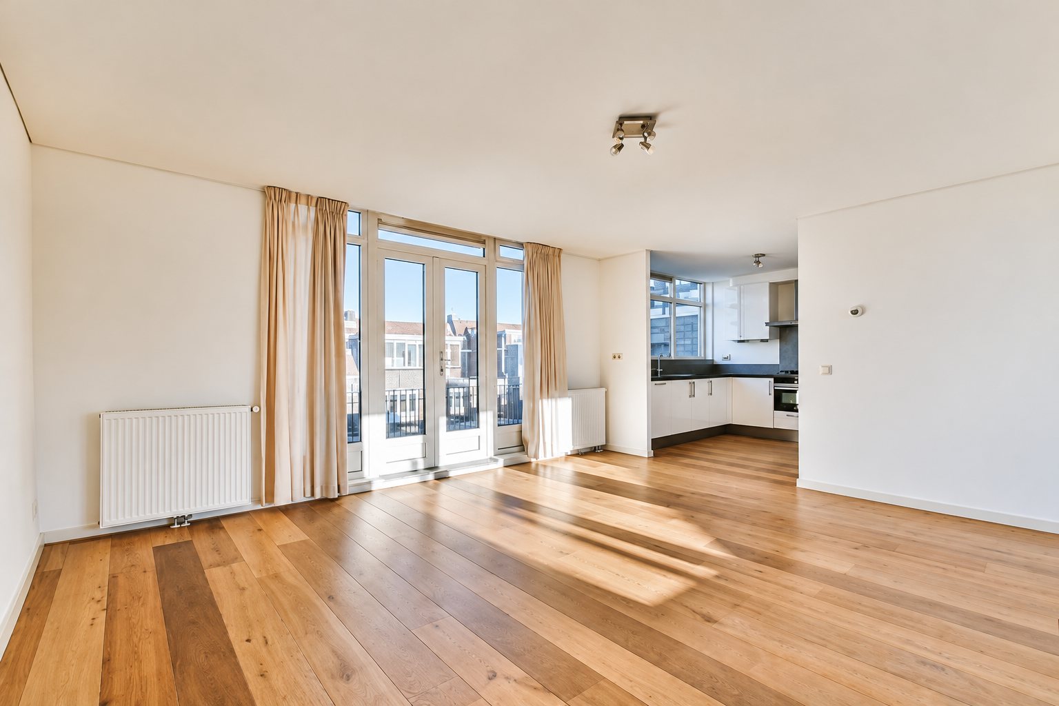 Un salon vide baigné de lumière avec un parquet en bois clair, de grandes portes-fenêtres aux rideaux beiges et une cuisine ouverte au fond.